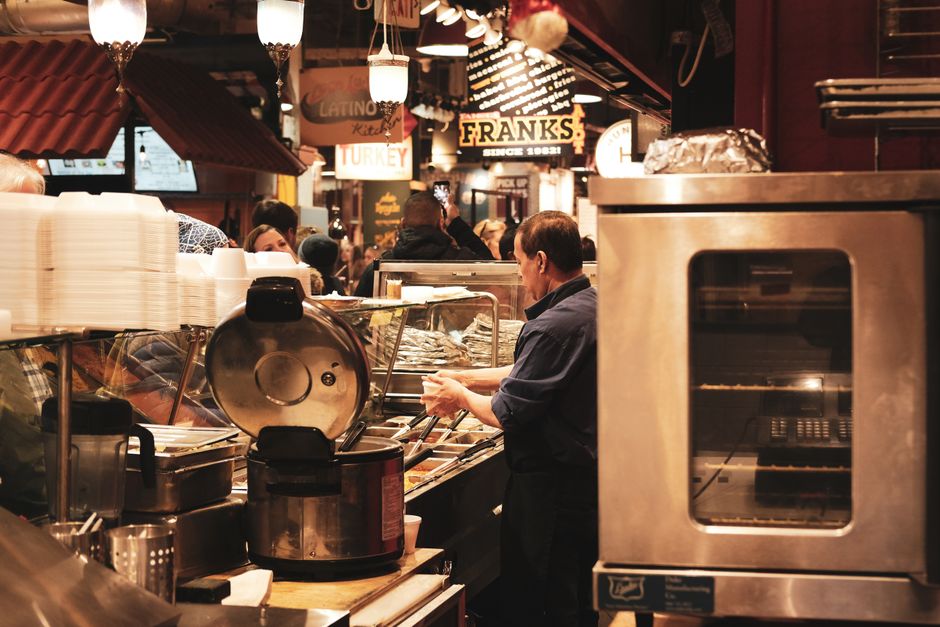 Inside a busy café on Edgware High Street, a staff member is preparing food behind a stainless steel counter with various cooking utensils, containers, and a rice cooker. The counter surface appears 