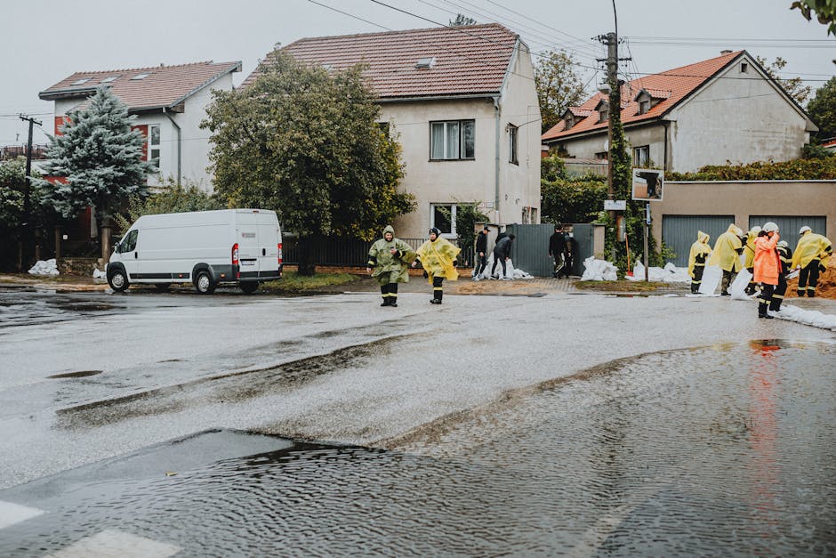 An outdoor scene showing a residential street with wet asphalt and standing water, likely from recent flooding or leaks. Several emergency workers dressed in yellow and green protective rain gear are