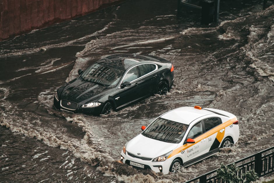 An outdoor scene showing a residential street with wet asphalt and standing water, likely from recent flooding or leaks. Several emergency workers dressed in yellow and green protective rain gear are